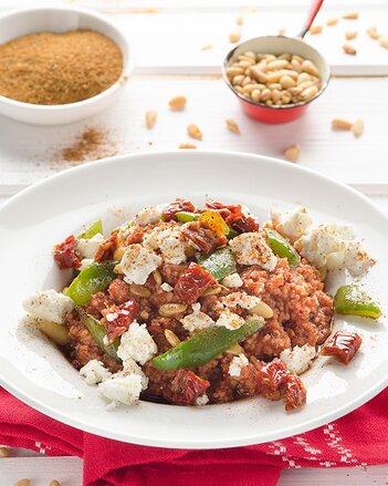 Auf dem Foto ist ein Teller mit Couscous-Tomaten-Pfanne zu sehen. Darüber ist Schafkäse verteilt. Im Hintergrund sieht man Pinienkerne. | © SONNENTOR