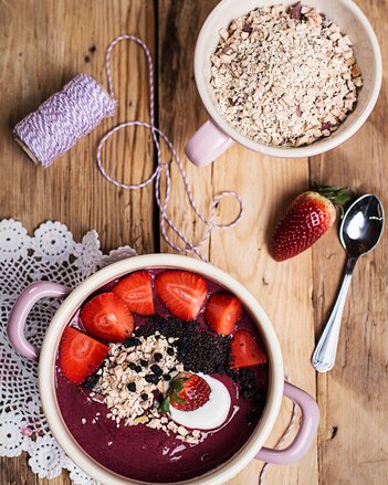 Foto einer Beeren Smoothie Bowl mit Superfood Topping. Daneben sieht man einen Löffel, eine Erdbeere und Haferflocken. | © SONNENTOR