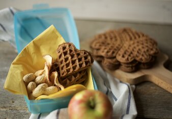 Auf dem Foto sind Schlaukakao Waffeln gemeinsam mit Erdnüssen in einer Jausenbox zu sehen. Im Hintergrund sieht man noch mehrere Waffeln. | © SONNENTOR