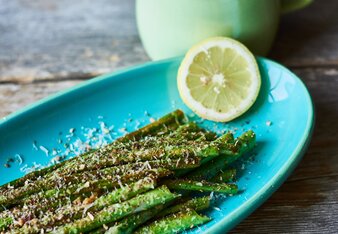 Auf dem Foto ist ein blauer Teller mit grünem Spargel darauf zu sehen. Der Spargel ist mit Parmesan bestreut, und daneben liegt eine Zitronenscheibe. | © SONNENTOR