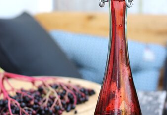 Auf dem Foto sieht man eine Flasche mit Hollerbalsamico. Daneben sieht man frische Holunderbeeren.  | © SONNENTOR