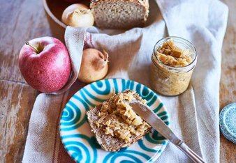 Auf dem Foto ist zu sehen, wie de Apfel-Zwiebel-Aufstrich auf ein Brot gestrichen wird. Daneben ist das Brot und das Glas mit Aufstrich zu sehen. | © SONNENTOR