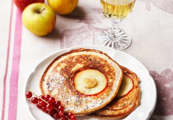 Auf dem Foto ist ein weißes Teller mit Crêpes auf einem rosa Tischtuch zu sehen. Dahinter sind Äpfel zu sehen. | © SONNENTOR