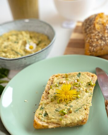 Ein Brötchen bestrichen mit dem Wildkäuterhummus und dekoriert mit einer Löwenzahnblüte liegt auf einem Teller, im Hintergrund steht die Schüssel mit dem restlichen Hummus. | © SONNENTOR