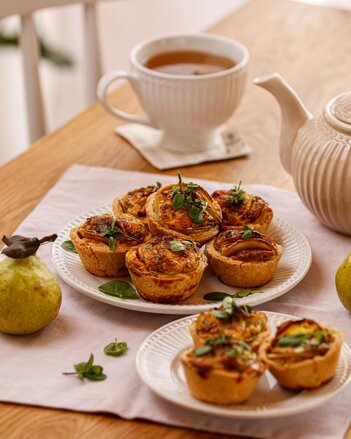 Die Mini Erdäpfel Quiches liegen auf 2 Tellern verteilt, dekoriert mit Basilikum und Majoran Blättern. Im Hintergrund steht eine Tasse Tee, 2 Birnen liegen daneben.  | © SONNENTOR