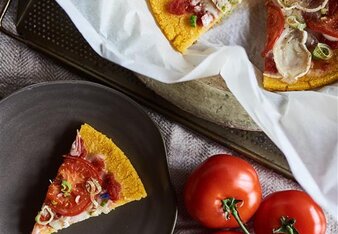 Auf dem Foto sieht man eine Polenta Pizza. Ein Stück wurde heraus genommen und liegt auf einem schwarzen Teller. Daneben sind Tomaten zu sehen. | © SONNENTOR