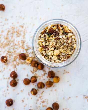 Auf dem Foto ist ein Glas mit dem Nuss-Gewürze-Topping zu sehen. Daneben liegen einige Haselnüsse und etwas von dem Topping. | © SONNENTOR