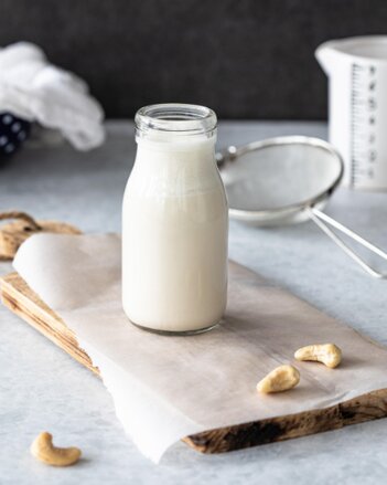 Glasflasche mit pflanzlicher Sahne steht auf einem Holzbrett. Darum herum ein paar Cashewnüsse. | © SONNENTOR