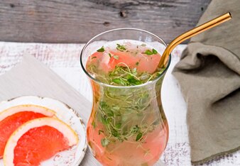 Ein bauchiges Cocktailglas ist gefüllt mit orange rosa Saft mit SONNENTOR Tschin Tschin Sirup, Grapefruit und Thymian, ergänzt mit einem rosegoldenen Trinkhalm. Ein Teller mit Grapefruit-Spalten steht daneben. | © SONNENTOR