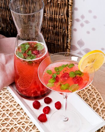 Ein Cocktailglas steht neben dem Krug, beides gefüllt mit einem roten Mocktail, gemacht aus Himbeeren und dem SONNENTOR Tschin Tschin Sirup. Kleine Minzblättchen schwimmen darin, das Glas ist mit einer Zitronenscheibe garniert. | © SONNENTOR