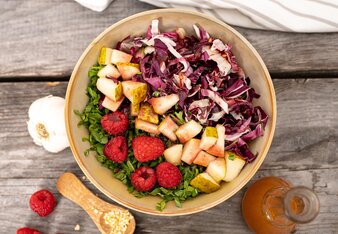 Ein Schüssel mit geschnittenem Rucola, Rotkraut und Birnen, mit Himbeeren am Rucola verteilt, steht zwischen einer Knoblauchknolle, einem Holzlöffel mit Knoblauchgranulat und 2 Dressingvasen auf einem Holztisch.  | © SONNENTOR