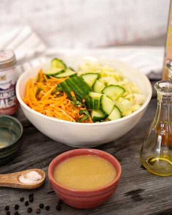 Die Salatschüssel ist gefüllt mit Karottenstiften, Gurkenstücken und Weißkraut und steht neben einer Dose Ayurvedischen Zaubersalz, einem Schälchen mit Dressing und einem Holzlöffel mit Salz. | © SONNENTOR