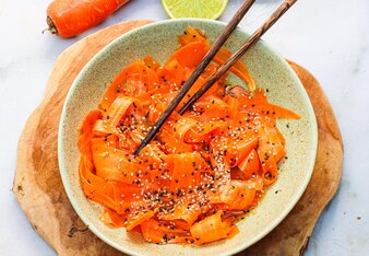 Der in Streifen geschnittene Karottensalat ist garniert mit Sesamkörnern und befindet sich in einer Schüssel, die auf einem Holzbrett steht. Asiatische Stäbchen stecken darin und daneben liegen noch einige Karotten sowie eine halbe Limette. | © SONNENTOR