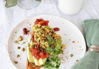 Das Frühstücksbrot ist mit Bohneneintopf, Eierspeis und Avocado belegt und mit Pistazien verziert und liegt auf einem Teller. Daneben steht eine Tasse Tee.  | © SONNENTOR