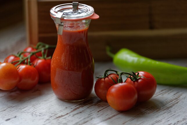 Auf dem Foto sieht man ein Glas voll mit Ketchup. Daneben sind frische Tomaten und einen grünen Paprika zu sehen. | © SONNENTOR