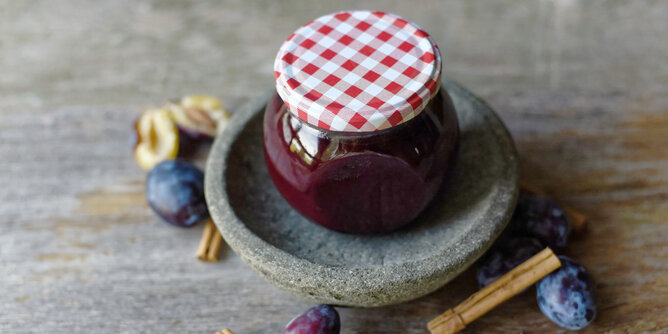 Auf dem Foto ist ein Glas Zwetschken Marmelade zu sehen. Daneben sieht man frische Zwetschken und Zimtstangen. | © SONNENTOR