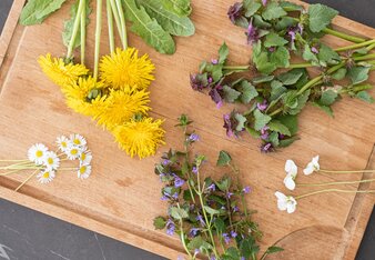 Auf dem Foto sieht man ein Holzbrett mit essbaren Blüten. Beispielsweise sieht man Gänseblümchen, Löwenzahn und Taubnessel. | © SONNENTOR